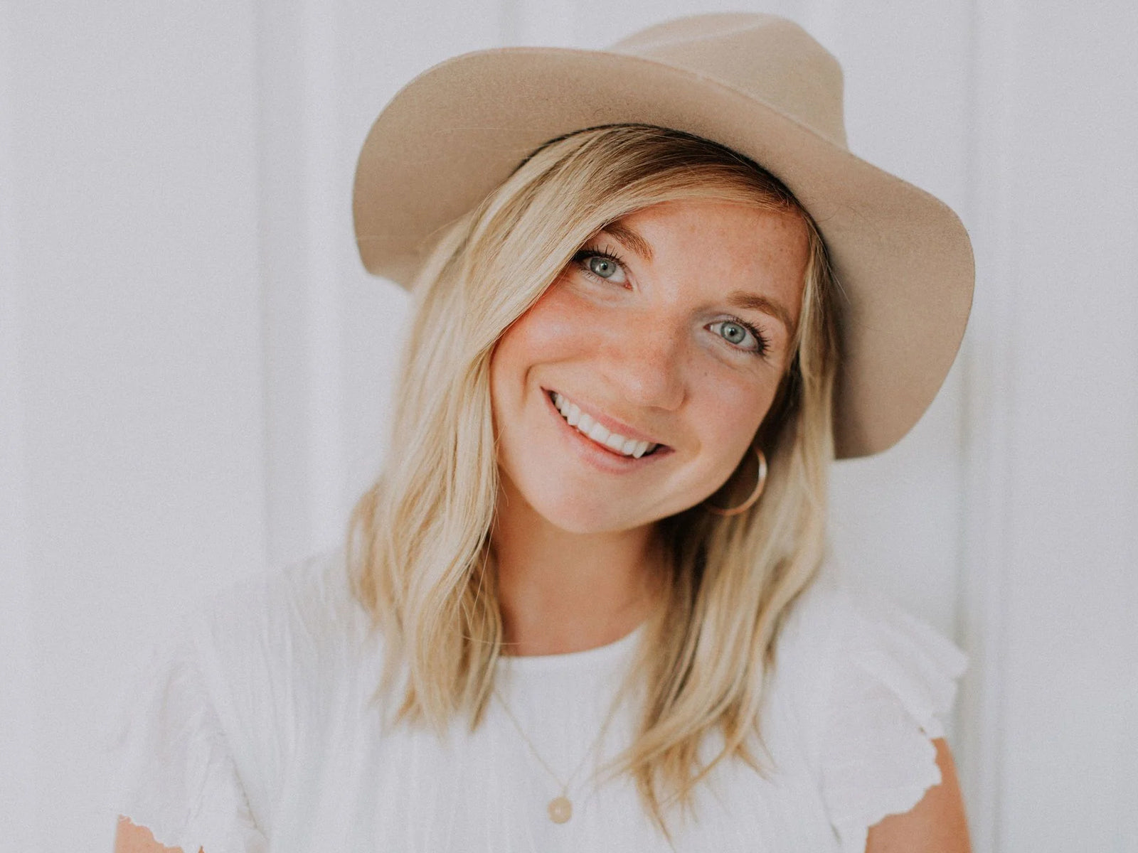 The founder of Hart + Stone wearing a beige wide-brim fedora hat and a white ruffled top, photographed against a soft white background.