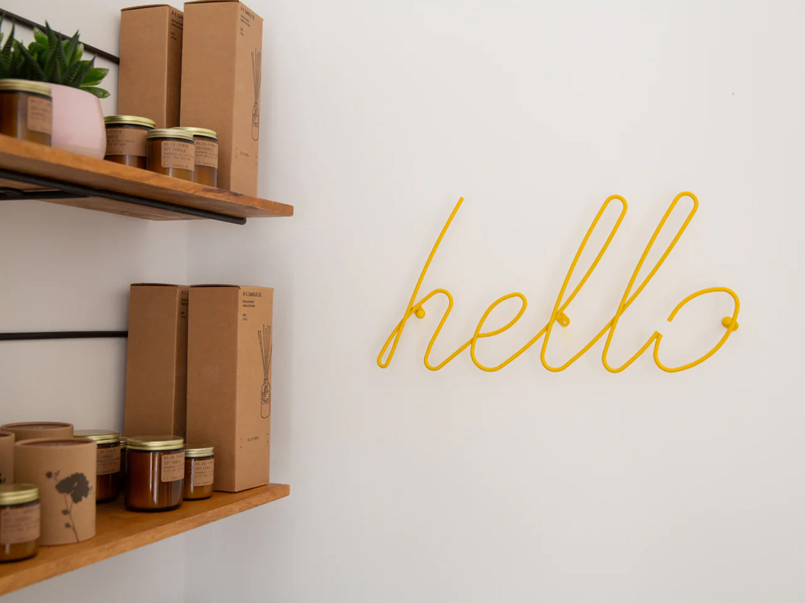 A yellow neon-style wire sign spelling 'hello' mounted on a white wall, with wooden shelves and small jars on the left side of the frame.
