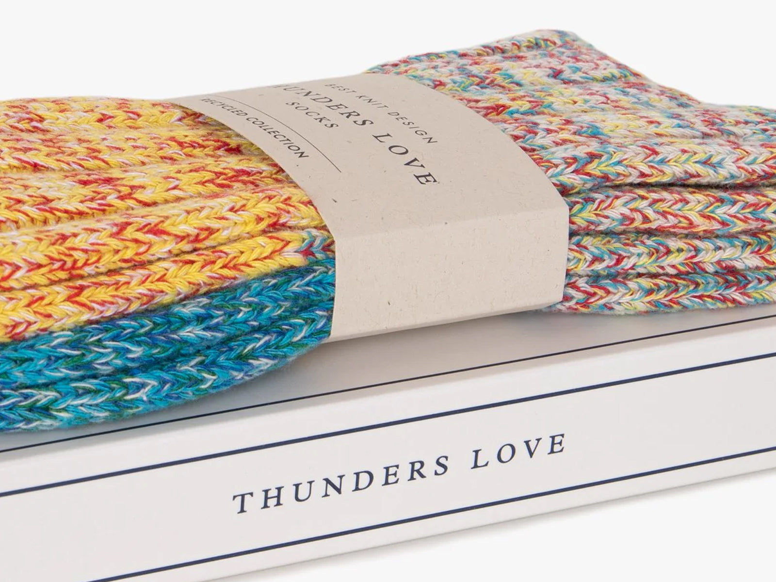 A close-up of folded multicolor knit socks wrapped with a beige paper band, resting on a white box printed with the brand name 'THUNDERS LOVE'.