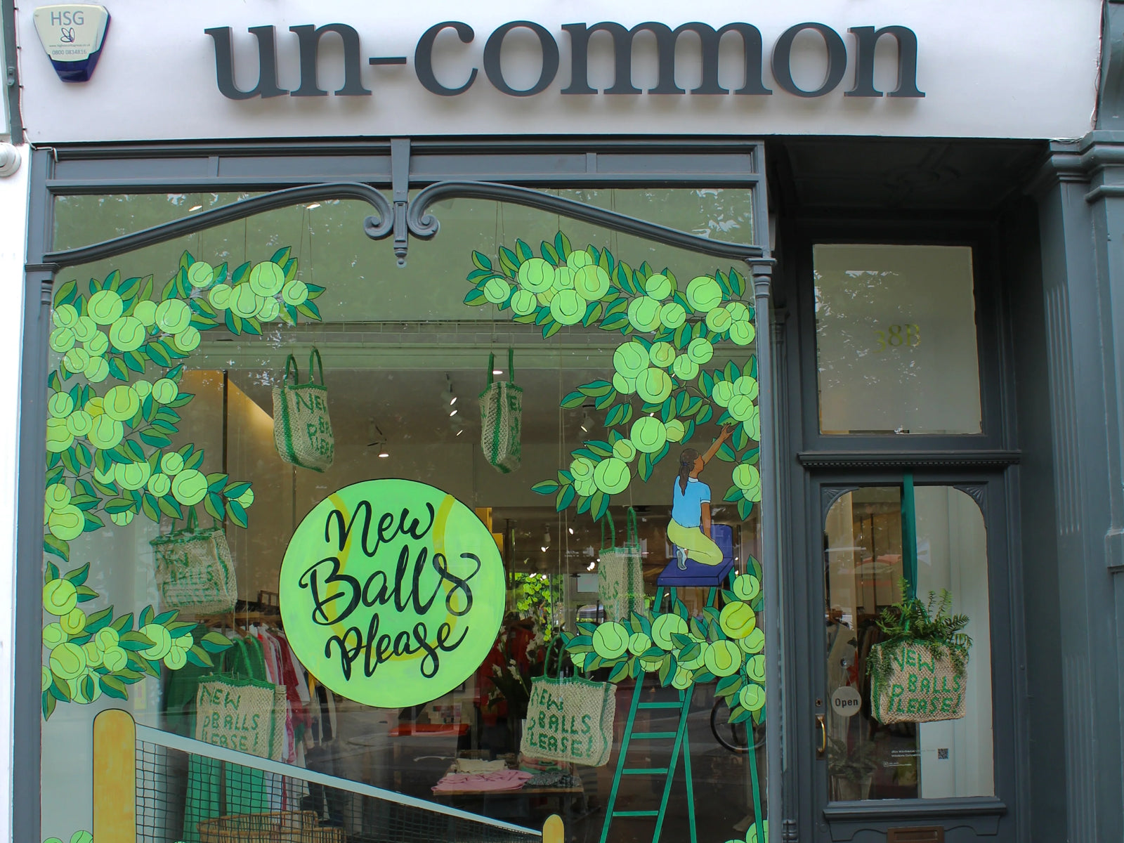 Un-Common storefront with a large window display decorated with green tennis ball graphics, a circular sign reading 'New Balls Please,' and a dark grey frame.