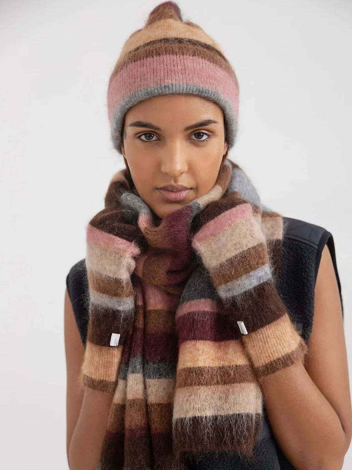 A model wearing a cozy knit beanie and matching striped scarf in earthy tones of brown, beige, pink, and grey.