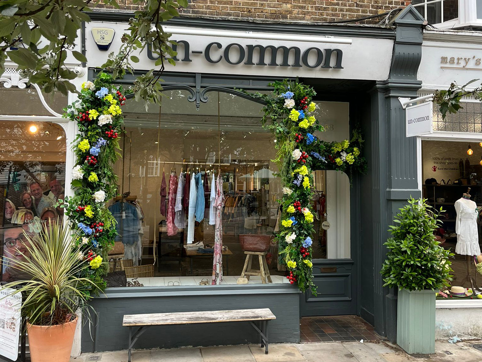 A storefront for Wimbledon based boutique un-common, featuring a dark grey facade, a wooden bench in front, and floral garlands framing the large glass windows.