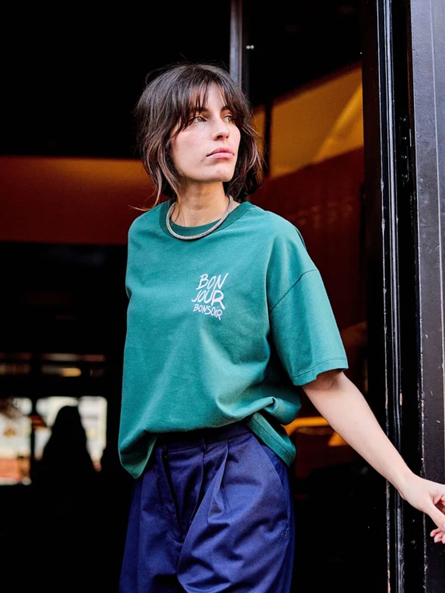 Woman wearing a green t-shirt saying 'Bon Jour' with blue trousers, in a doorway.