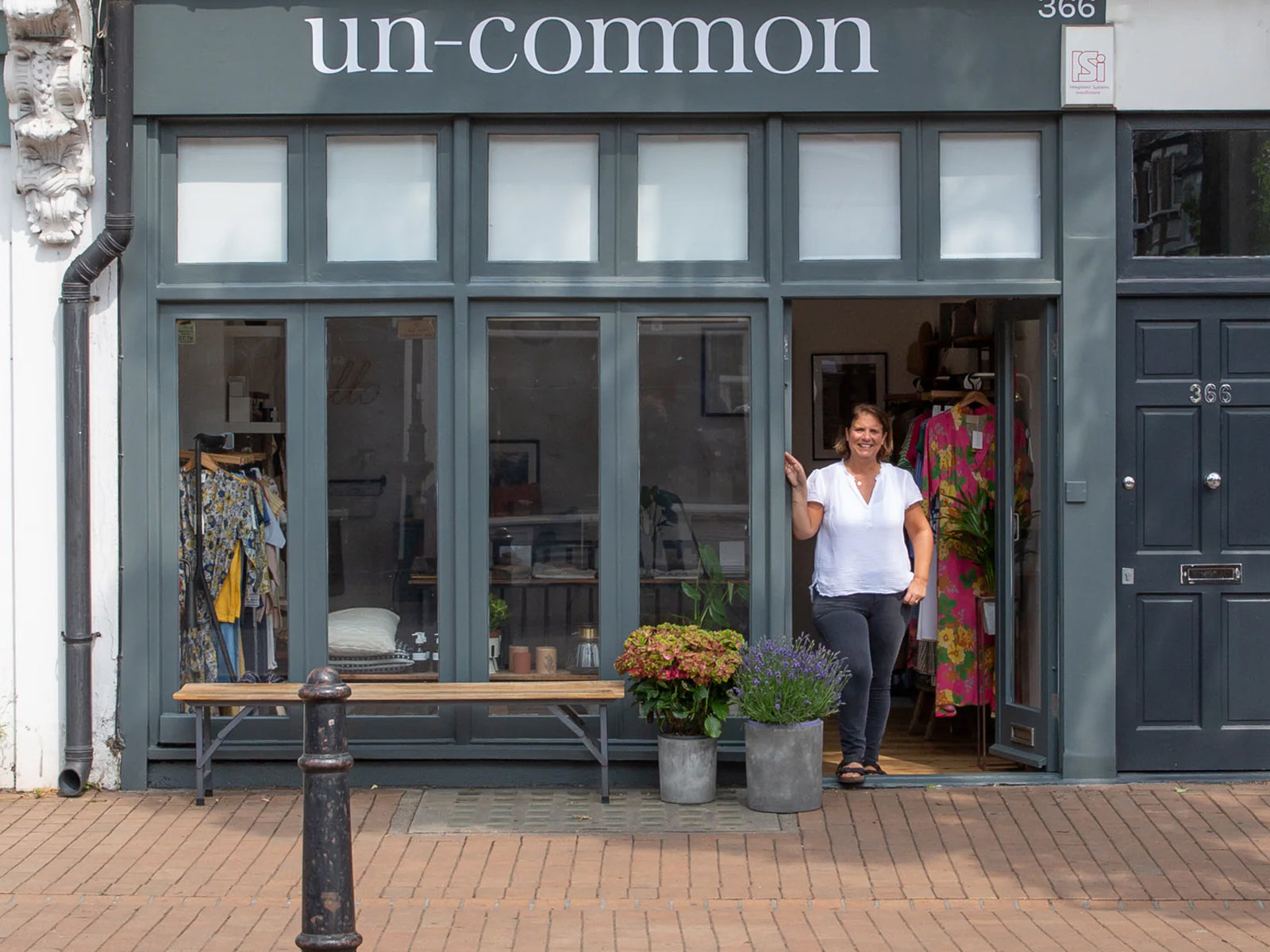 Kate stands in the doorway of the Un-Common storefront with the sign 'un-common' above large dark grey windows. The shop interior shows clothing racks with colourful garments.